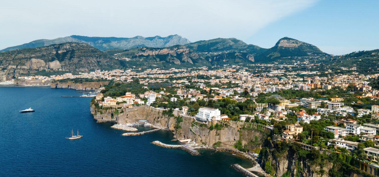 Aerial drone view of the Tyrrhenian sea coast in Sorrento, Italy. Multiple classic buildings on rocky cliffs, a lot of greenery and hills in the distance