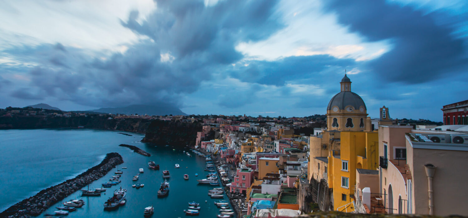 View of the Port of Corricella with lots of colorful houses after sunset in Procida Island, Italy.